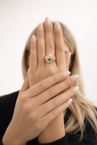 Kadın elinde takılı mavi taş detaylı göz motifli VIP gold yüzük, nude pembe manikürlü bakımlı el, beyaz stüdyo arka plan, lüks mücevher ürün fotoğrafı.