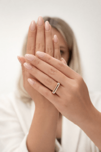 Bezel taş detaylı altın yüzük takan zarif kadın eli, nude pembe manikürlü, açık bej stüdyo arka planında lüks ürün çekimi